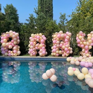 May include: A large "BABY" sign made of gold frames filled with pink and cream balloons. The sign is in front of a swimming pool.