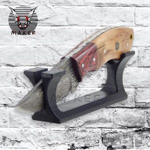 May include: A handcrafted knife with a patterned blade and a wooden handle with red accents. The knife rests on a black display stand against a white brick wall. The word "MAKER" is visible.