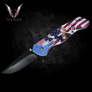 May include: A black folding knife with a blue and red American flag design. The handle features the words "FIGHT FIGHT FIGHT" printed on it. The blade is partially open.