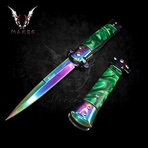 Op de afbeelding: Een regenboogkleurig Milano stiletto mes met een groen gemarmerd handvat. Het mes is open en het lemmet reflecteert verschillende kleuren. Het handvat is levendig groen met een gemarmerd patroon. Het mes staat tegen een donkere achtergrond.