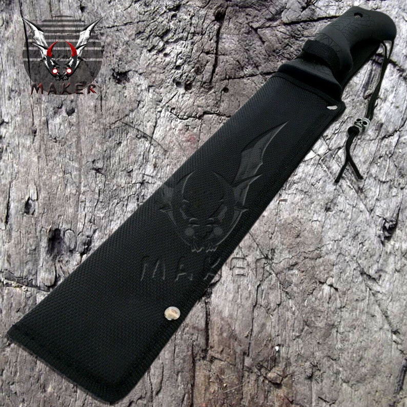 Op de afbeelding: Zwarte machete met een zwarte schede. De schede heeft een schedel- en vleermuisontwerp met het woord "MAKER" erop gegraveerd.
