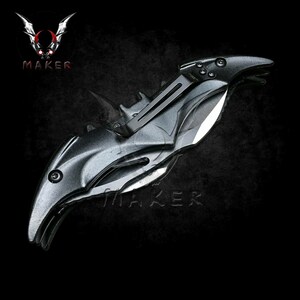 Puede incluir: Un cuchillo plegable negro con forma de murci&eacute;lago con una hoja plateada. El cuchillo tiene un mango negro texturizado y un mecanismo de resorte.