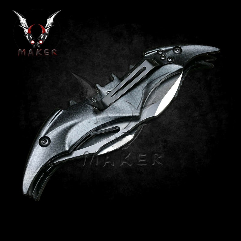 Puede incluir: Un cuchillo plegable negro con forma de murci&eacute;lago con una hoja plateada. El cuchillo tiene un mango negro texturizado y un mecanismo de resorte.