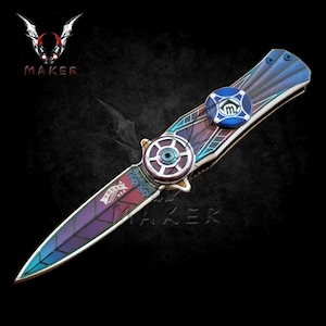 May include: A purple, blue, and silver folding knife with a decorative design. The knife has a spinning wheel on the handle and a blue and silver emblem with the letter "M" on the side.