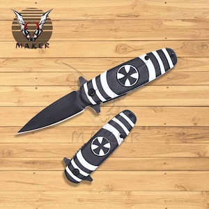 May include: A black and white pocket knife with a cross design on the handle. The knife is open and the blade is visible.