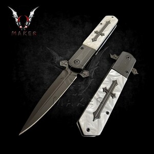 May include: A black and white folding knife with a cross design on the handle. The blade is black and has a sharp point. The knife is closed and the handle is visible.
