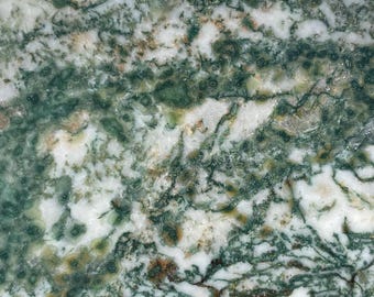 May include: A polished, irregularly shaped stone with a striking pattern of green, white, and brown veining. The stone is set against a backdrop of rough, textured rock and weathered wood, showcasing its natural beauty.