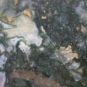 May include: A square-shaped moss agate stone with a natural, organic pattern. The stone features a mix of dark green, white, and tan colors, creating a unique, textured appearance. The stone is held by a person's fingers.