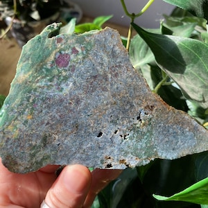 May include: A large, flat, green and gray stone with a rough, irregular surface. The stone has a few small holes and is speckled with red and yellow.