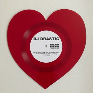 Op de afbeelding: Een rode hartvormige vinylplaat met een wit label waarop "DJ DRASTIC" en "WNW RECORDINGS" staat. Het label bevat ook de titel van het nummer "CHE NELLE FEAT. CHAM - I FELL IN LOVE WITH THE DJ [DJ DRASTIC DUBPLATE SPECIAL VERSION]" en de trackinformatie "01. MAIN [2:06]".