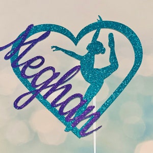 May include: A teal glitter heart with a silhouette of a dancer inside. The name "Meghan" is written in purple glitter on the left side of the heart.