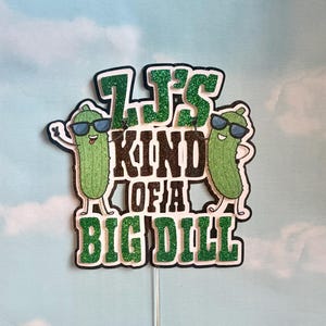 Puede incluir: Adorno para pastel con la frase "It's Kind of a Big Dill" en letras negras y purpurina verde. Dos pepinillos de dibujos animados con gafas de sol flanquean el texto, a&ntilde;adiendo un toque divertido al dise&ntilde;o.
