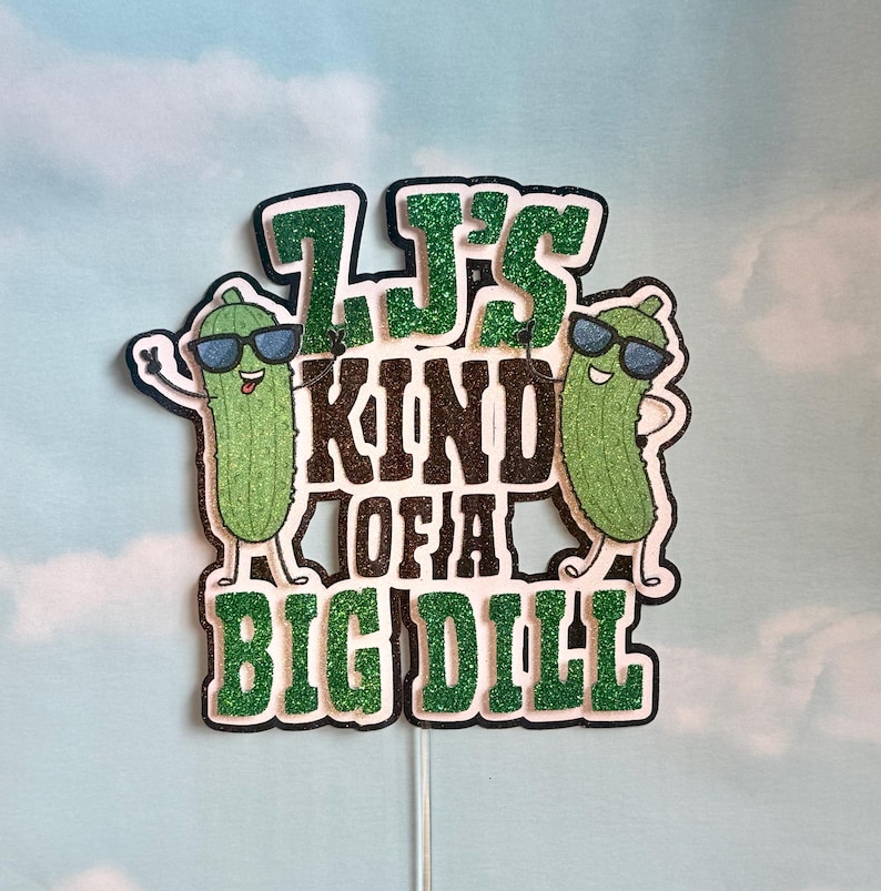 Puede incluir: Adorno para pastel con la frase "It's Kind of a Big Dill" en letras negras y purpurina verde. Dos pepinillos de dibujos animados con gafas de sol flanquean el texto, a&ntilde;adiendo un toque divertido al dise&ntilde;o.