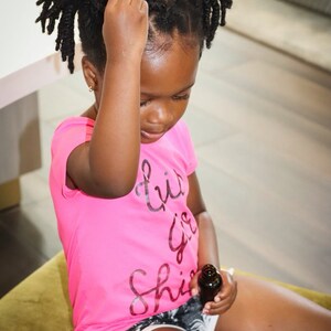 May include: A young person with dark brown hair styled in two braids sits on a green cushion and holds a small brown bottle. They are wearing a pink shirt with the text "Little Girl Shine" printed on it and black and white shorts.