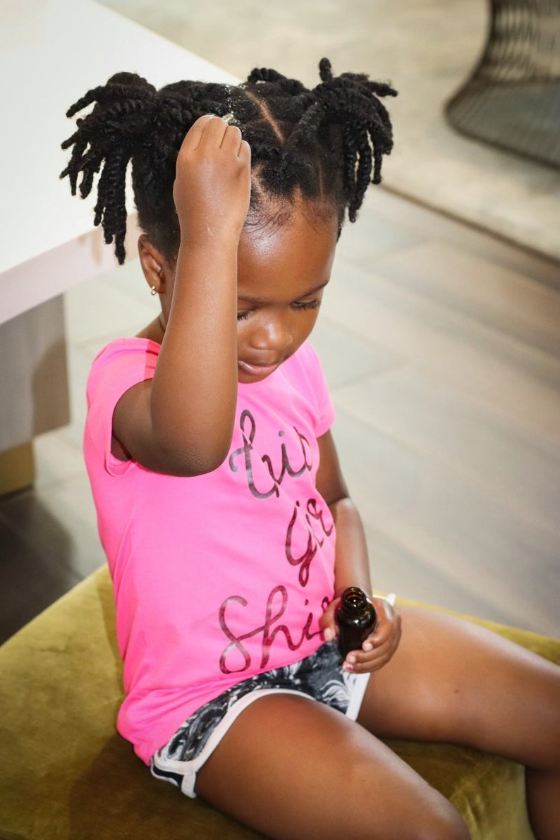 May include: A young person with dark brown hair styled in two braids sits on a green cushion and holds a small brown bottle. They are wearing a pink shirt with the text "Little Girl Shine" printed on it and black and white shorts.