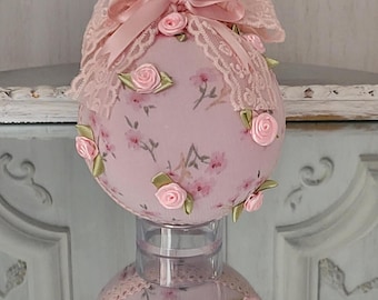 May include: A pink fabric Christmas ornament with a floral pattern and pink satin ribbon bow. The ornament is decorated with pink roses and green leaves.