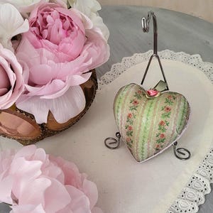 May include: A green and white heart-shaped pillow with a floral pattern and a pink rose on a white lace doily. The pillow is on a metal stand.