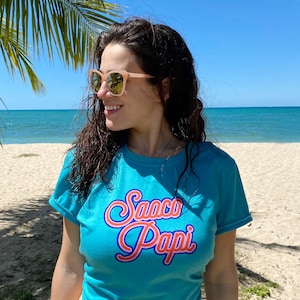 Könnte beinhalten: Eine Frau trägt ein türkisfarbenes T-Shirt mit dem Text "Saco Papi" in rosa und blauen Buchstaben. Sie trägt eine Sonnenbrille und steht am Strand.
