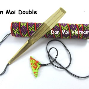 May include: A brass Dan Moi double-lip harp with a black string and a decorative case. The case is cylindrical and features a geometric pattern in red, purple, and green. The text "Dan Moi Double" and "Dan Moi Vietnam" are visible.