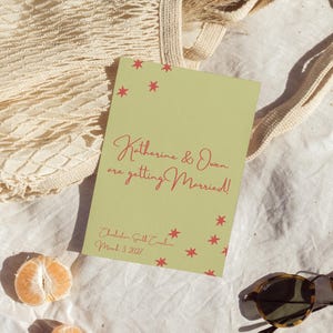 May include: A wedding invitation with the text "Katherine & Owen are getting Married!" in red script on a light green background. The invitation is surrounded by a beige net bag, sunglasses, and dried pampas grass.