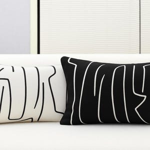 May include: Two rectangular throw pillows. One is black with a white abstract design, and the other is white with a black abstract design. The pillows are on a white surface, with a neutral background.