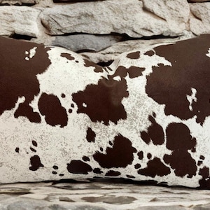 May include: A brown and white cowhide patterned throw pillow. The pillow is made of a soft, plush fabric and has a subtle sheen.