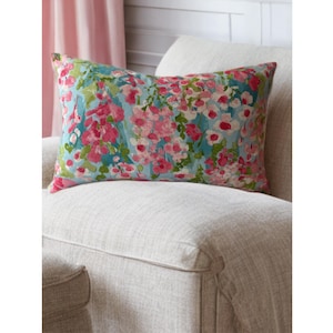 May include: Rectangular decorative pillow with a floral pattern. The pillow features a light blue background with pink and white flowers and green leaves. The pillow is placed on a beige upholstered chair.