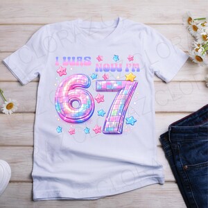 Pu&ograve; includere: T-shirt bianca con la scritta "I WAS NOW I'M" sopra i numeri "67". I numeri sono disegnati con un effetto palla da discoteca, circondati da stelle rosa, blu e gialle. Una divertente maglietta di compleanno.