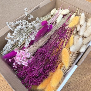 Pode incluir: Uma caixa de cartão preenchida com uma variedade de flores secas. O arranjo inclui flores brancas, rosas e roxas, juntamente com gramíneas amarelas e bege. As flores são dispostas em feixes, criando uma exibição colorida e texturizada.