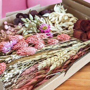 May include: A box filled with dried flowers and grasses in shades of pink, white, and brown. The box is made of cardboard and has a natural brown color.
