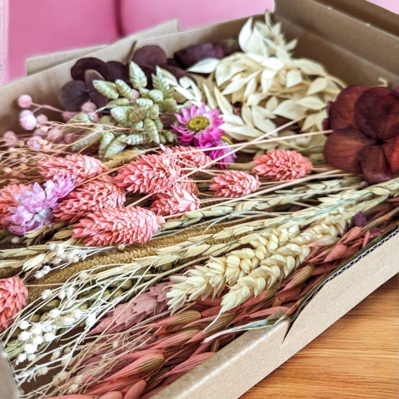 May include: A box filled with dried flowers and grasses in shades of pink, white, and brown. The box is made of cardboard and has a natural brown color.