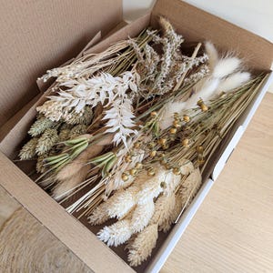 Könnte beinhalten: Eine Schachtel gefüllt mit getrockneten Blumen und Gräsern in Weiß-, Beige- und Grüntönen. Die Schachtel ist geöffnet und die Blumen fallen heraus.
