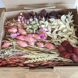 May include: A box filled with a variety of dried flowers and foliage, including pink, white, and burgundy shades. The flowers are arranged in a bouquet-like fashion, with some stems extending beyond the box.