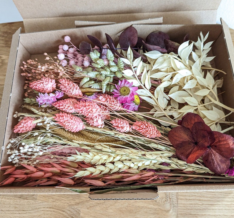 May include: A box filled with a variety of dried flowers and foliage, including pink, white, and burgundy shades. The flowers are arranged in a bouquet-like fashion, with some stems extending beyond the box.