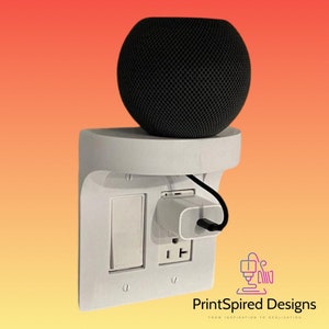 May include: A white 3D printed wall mount for a smart speaker, with a built-in outlet and switch. The mount is designed to hold a round black speaker, and the speaker is plugged into the outlet. The text "PrintSpired Designs" is visible in the bottom right corner of the image.