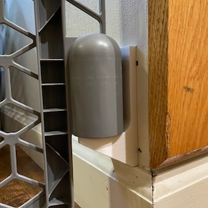 May include: A gray baby gate with a white plastic mounting bracket attached to a wall. The gate is made of metal and has a diamond pattern.