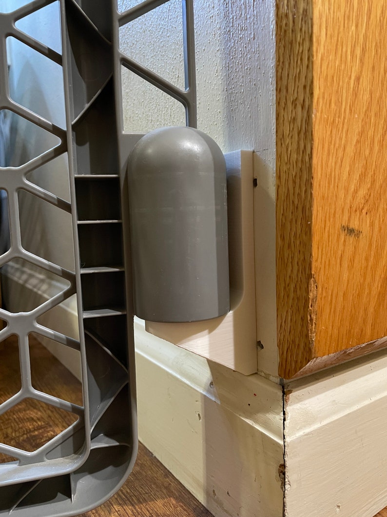 May include: A gray baby gate with a white plastic mounting bracket attached to a wall. The gate is made of metal and has a diamond pattern.