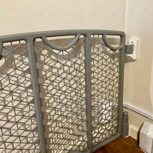 May include: A gray plastic baby gate with a diamond-shaped mesh pattern. The gate is attached to a white wall with a white trim.