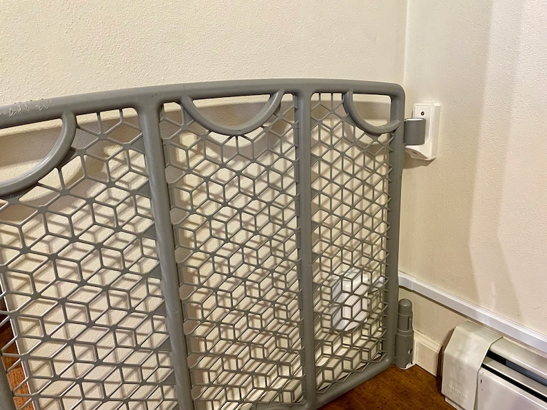 May include: A gray plastic baby gate with a diamond-shaped mesh pattern. The gate is attached to a white wall with a white trim.