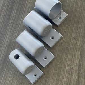 May include: Four gray plastic wall mounts with different sized holes. The mounts are designed to hold pipes or rods.