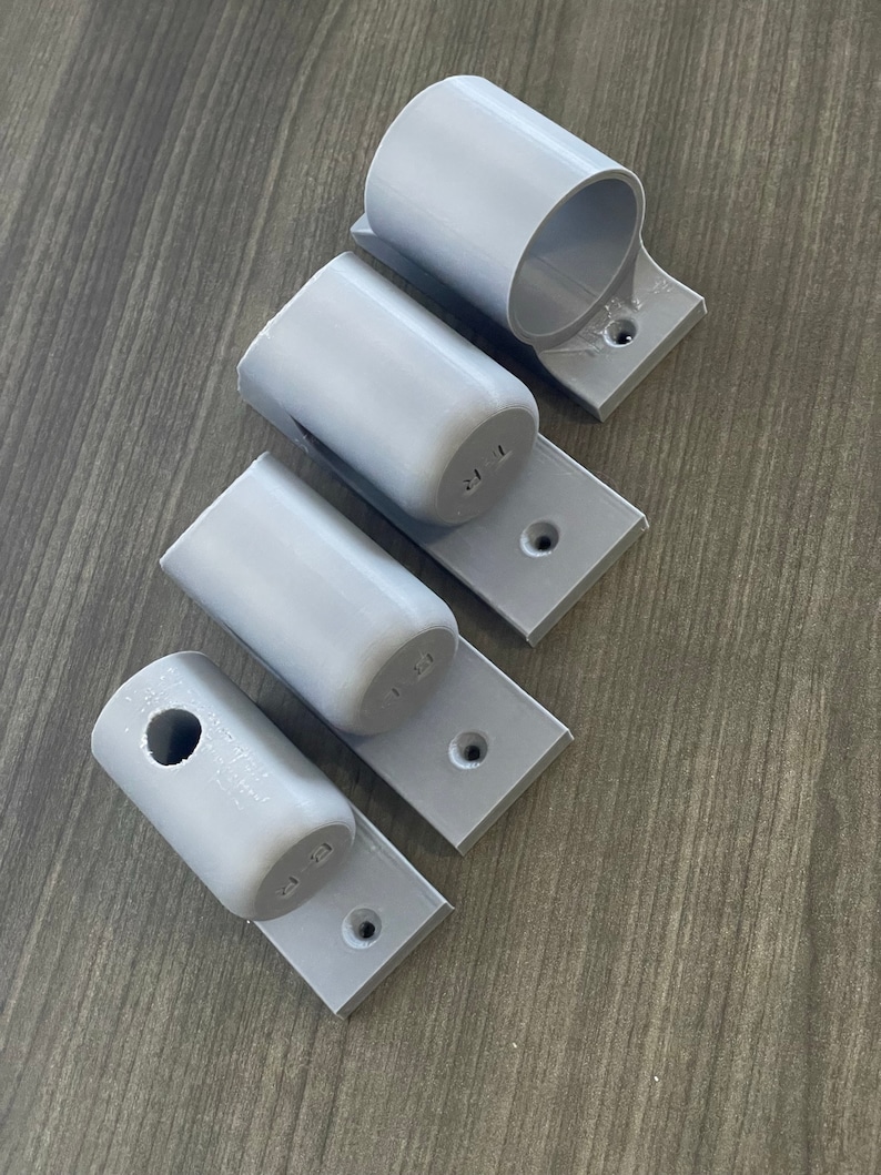 May include: Four gray plastic wall mounts with different sized holes. The mounts are designed to hold pipes or rods.