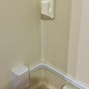 May include: White plastic electrical outlet covers and cord covers installed in a corner of a room. The cord covers are attached to the wall with white plastic clips.