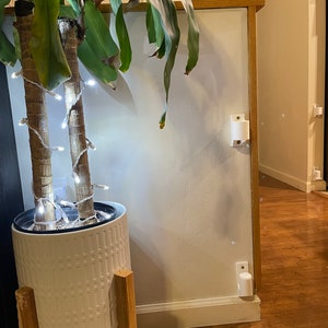 May include: A white planter with a textured pattern sits on a wooden stand. The planter is filled with a green plant with white string lights wrapped around the stems. The plant is in front of a white wall with a white trim.