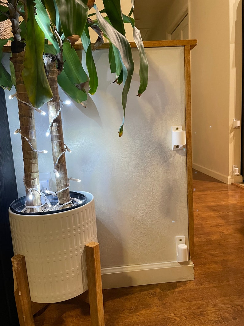 May include: A white planter with a textured pattern sits on a wooden stand. The planter is filled with a green plant with white string lights wrapped around the stems. The plant is in front of a white wall with a white trim.