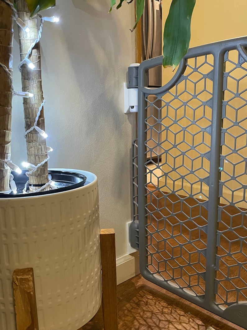 May include: A white planter with a patterned design sits on a wooden stand. The planter is filled with a green plant and white string lights. A gray baby gate is in the background.
