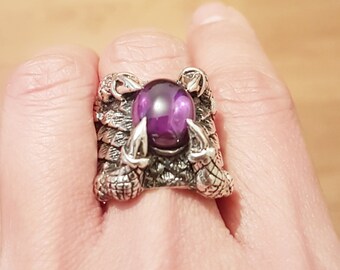 May include: A silver ring featuring a purple gemstone set in a claw design. The ring is detailed with intricate carvings and has a textured finish.