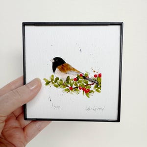 May include: A framed watercolor painting of a bird with a black head, brown back, and white belly, perched on a branch with green leaves and red berries. The artwork is signed "Lisa Livory" and numbered 1/500.