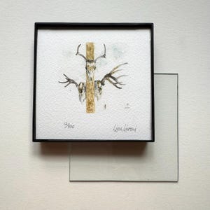 May include: A framed watercolor painting featuring deer skulls and antlers, centered on a vertical wooden post. The artwork is signed "Lisa Livoni" and numbered "12/500". The frame is black, and a glass pane is partially visible.