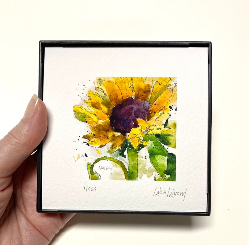 May include: A watercolor painting of a sunflower with yellow petals and a dark purple center. The painting is framed in a black frame and has the artist's signature and the number 1/500 in the bottom corner.