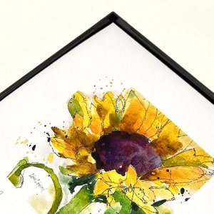 May include: A watercolor painting of a sunflower with yellow petals and a dark purple center. The painting is framed in a black frame and has the artist's signature "Lisa Livoni" and the number "1500" in the bottom left corner.
